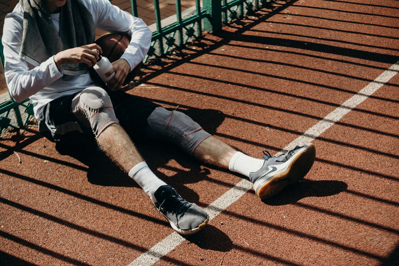 Young male athlete sitting on sports court holding a basketball and water bottle, knees bent.